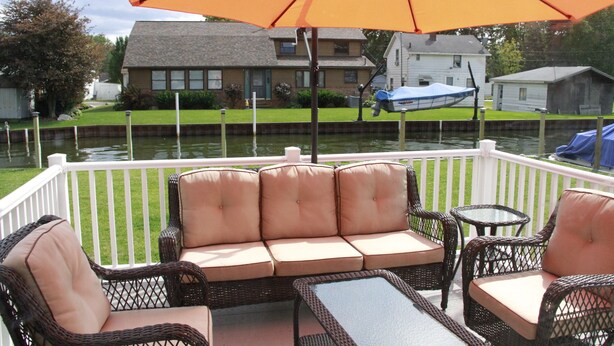 Une terrasse dotée de meubles en osier, d’un parasol et offrant une vue sur une maison et sur l’eau.