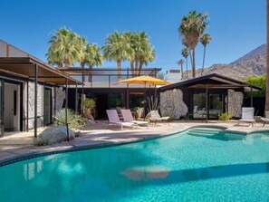 A modern pool area with lounge chairs and a covered seating area, surrounded by palm trees and a mountain backdrop.