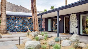 Un edificio moderno con un gran mural, palmeras y un jardín desértico con rocas y cactus.