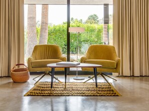 A room with two yellow armchairs, a small table, a lamp, a woven basket, and a view of greenery through large windows.