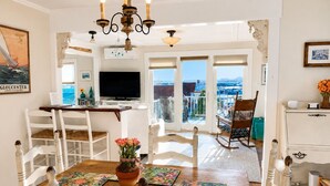 A dining area with a wooden table, chairs, a chandelier, and a view through glass doors to the outside. - Romantic Summerwind Cottage-Harbor View Retreat w/Large Private Deck (Gloucester)