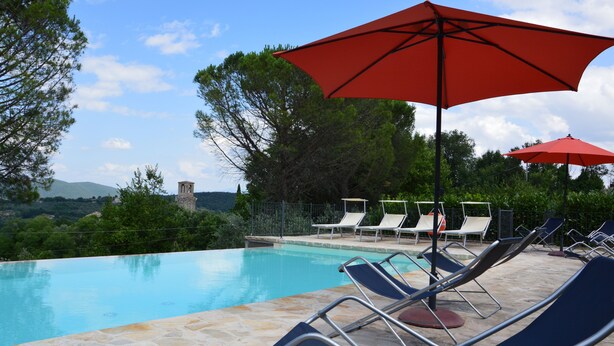 Zona de piscina con sillas de descanso y sombrillas rojas, rodeada de árboles y vistas a montañas distantes.