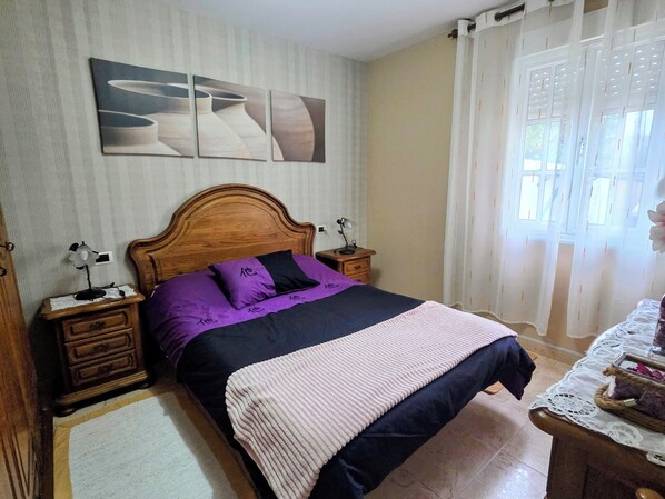 A bedroom with a wooden headboard, a bed with a black and purple bedspread, two bedside tables with lamps, a wall-mounted artwork, and a window with curtains.