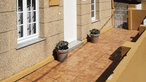 Un edificio con fachada de piedra, una puerta blanca y un pequeño ventanal con contraventanas blancas. Hay dos macetas con plantas frente a la puerta.