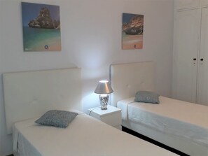 Quarto de hotel com duas camas de solteiro, mesa de cabeceira com abajur, e duas imagens emolduradas de uma praia na parede.