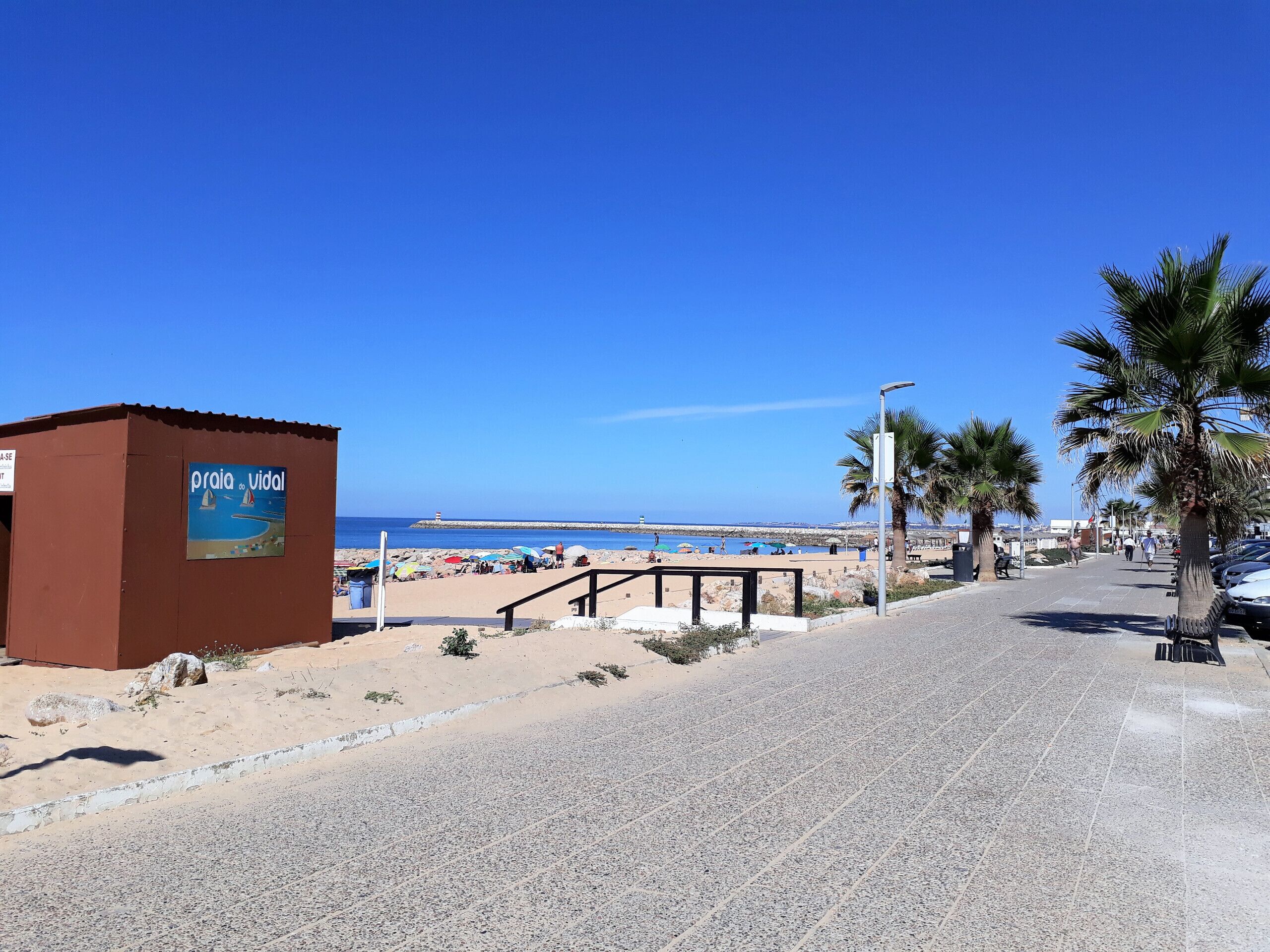 Een strandboulevard met een bord waarop "Praia do Vial" staat, een verharde looppad, palmbomen en de zee op de achtergrond.
