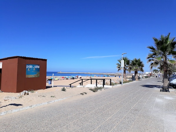 Um calçadão à beira-mar com uma placa indicando 'Praia do Vial', um passeio pavimentado, palmeiras e o mar ao fundo.