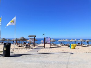 Una playa con sombrillas, un cartel y un contenedor de basura.