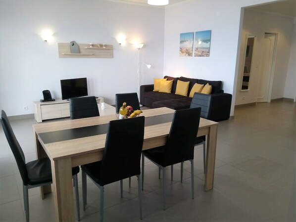 Uma sala de estar moderna com área de jantar, apresentando uma mesa de jantar de madeira, cadeiras pretas, um sofá com almofadas amarelas e uma TV de tela plana sobre um suporte de madeira.