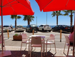 Um café à beira-mar com mesas ao ar livre e guarda-sóis vermelhos.