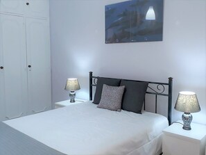 Una cama bien tendida con dos almohadas, una mesita de noche con una lámpara y un cuadro colgado en la pared.