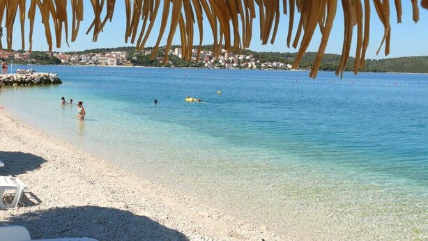 A beach with clear water, a thatched roof structure, and a distant town.