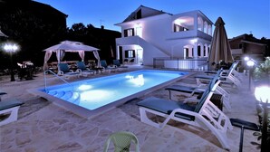A swimming pool with lounge chairs and umbrellas, surrounded by a stone patio and a two-story building with a balcony.