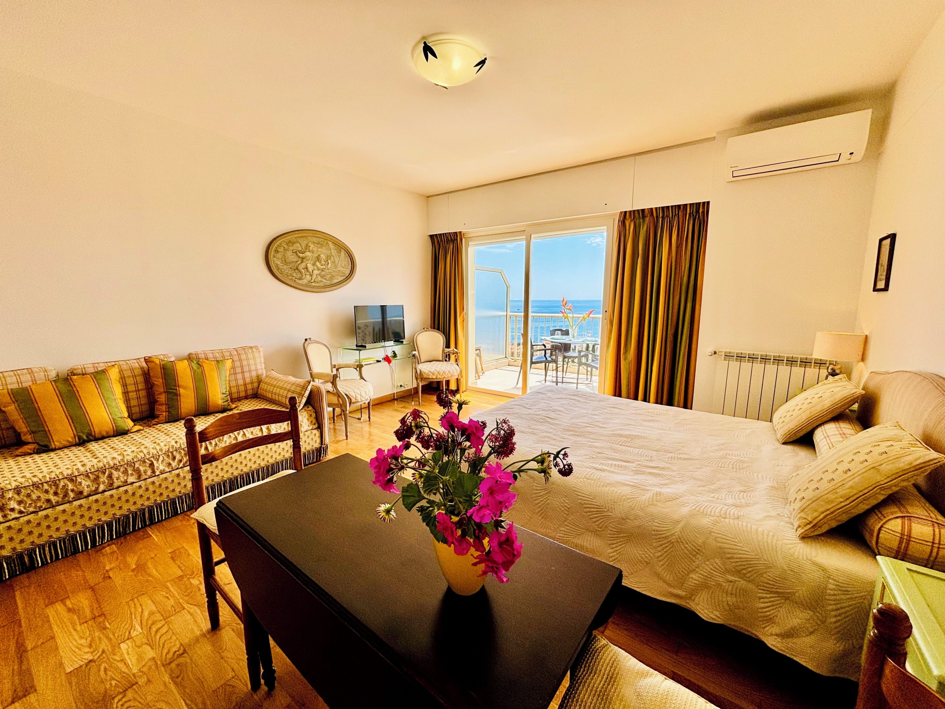 A hotel room with a bed, a sofa, a TV, a table with flowers, and a balcony with a view.