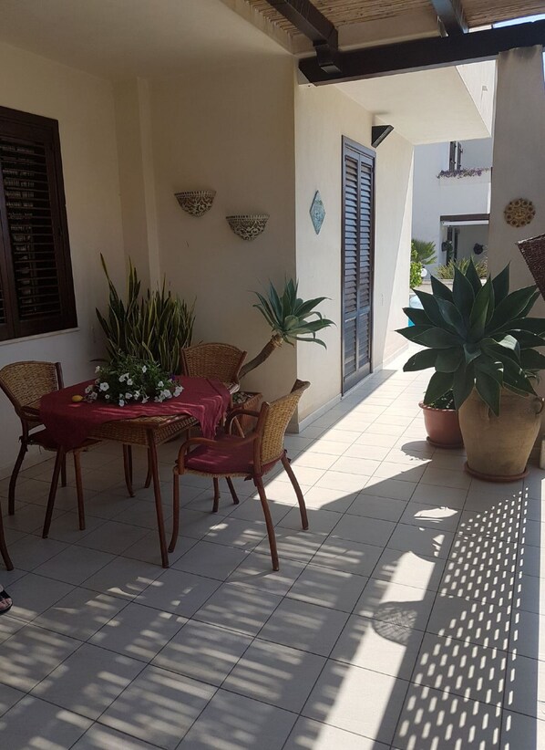An outdoor patio with a table and chairs, surrounded by potted plants and decorative wall hangings.