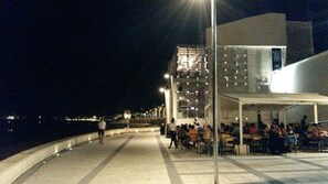 A waterfront promenade with outdoor seating, a modern building with illuminated windows, and a view of the sea.