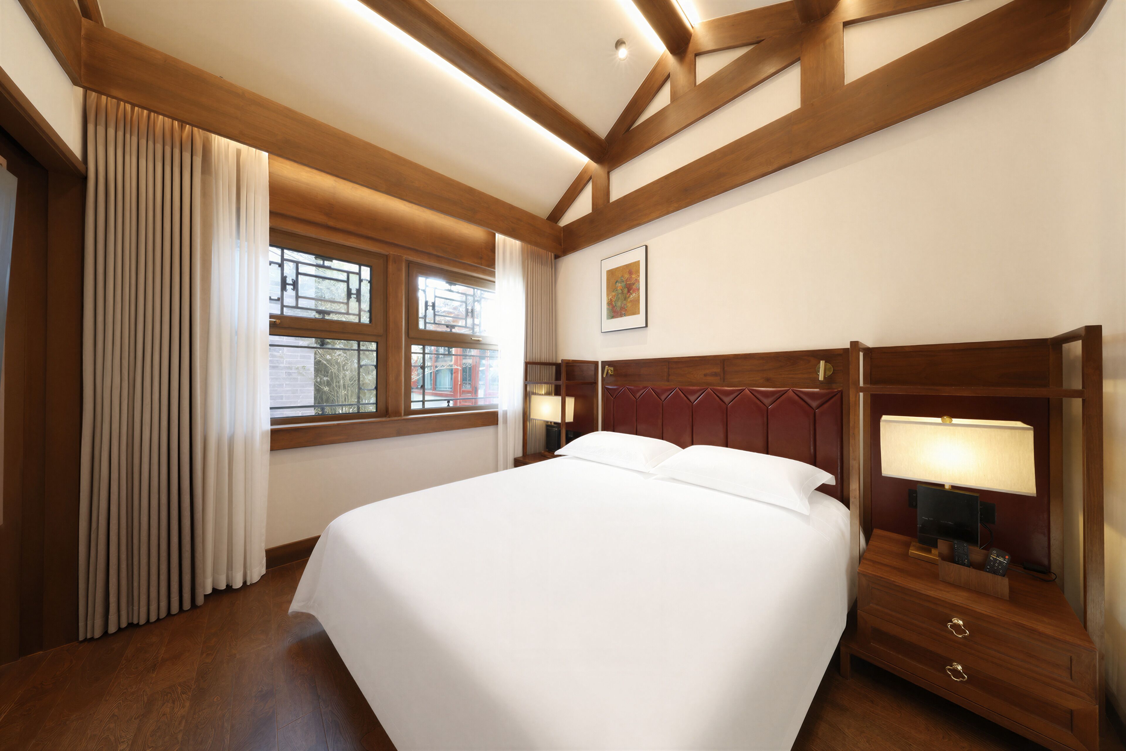 A hotel room with a large bed, wooden headboard, bedside lamps, a window with curtains, and a framed picture on the wall.
