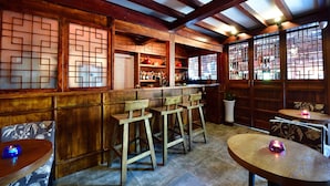 Uma área de bar com banquetas de madeira, balcão e prateleiras com garrafas. Há duas mesas com cadeiras, e uma planta em vaso é visível.