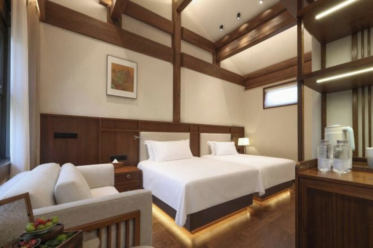 A hotel room with two beds, a wooden headboard, a bedside table with a lamp, a small seating area with a chair, and a fruit bowl.