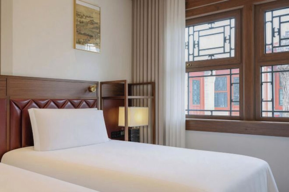 A hotel room with a bed, a nightstand, a lamp, and a window with a view of buildings.