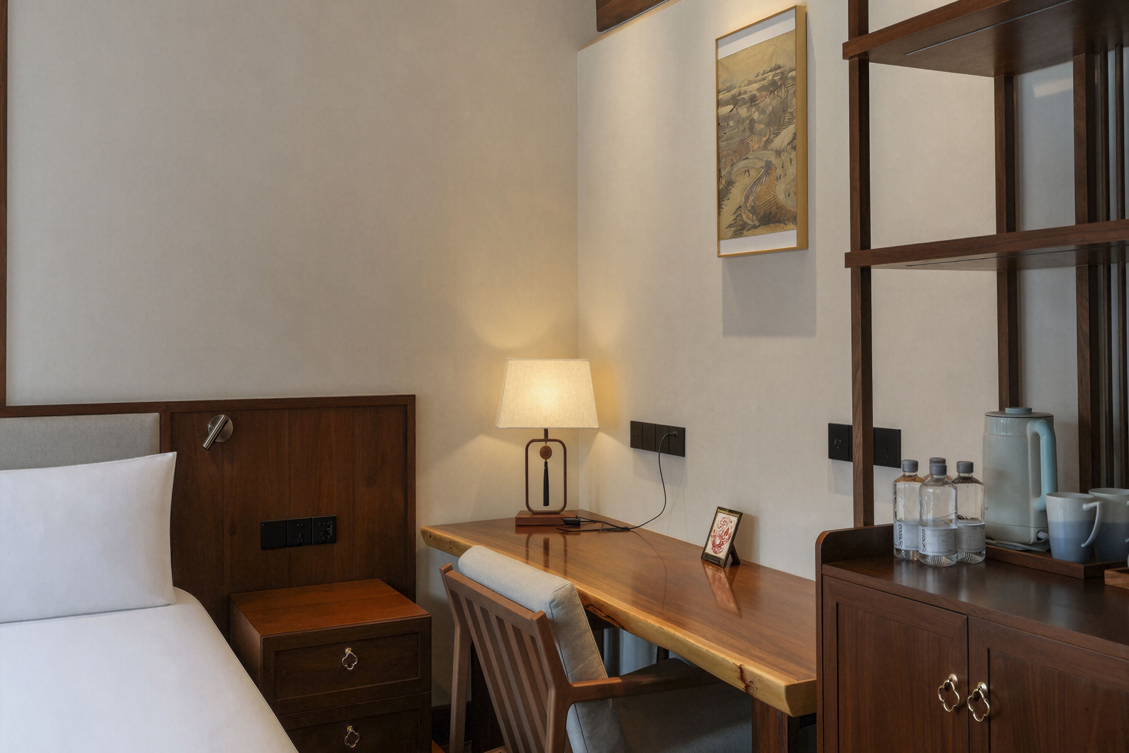 A hotel room with a bed, a desk, a chair, a lamp, a picture frame, a cabinet, and a coffee maker.