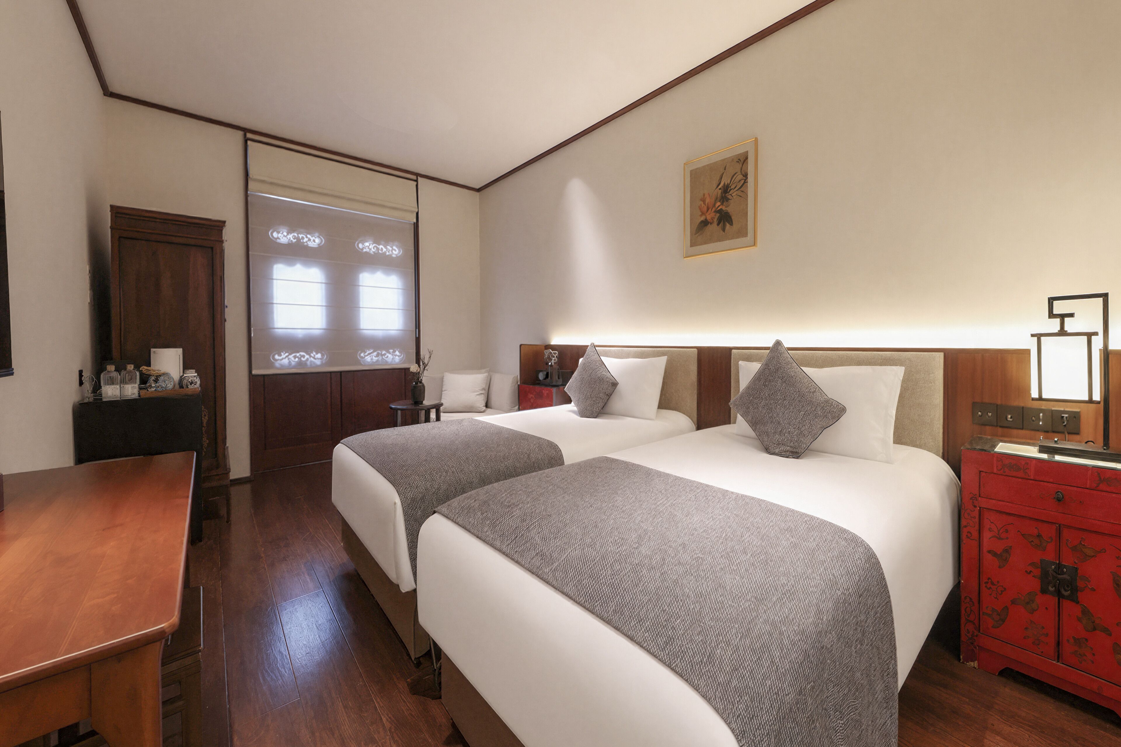 A hotel room with two beds, a wooden desk, a dresser, and a window with blinds.