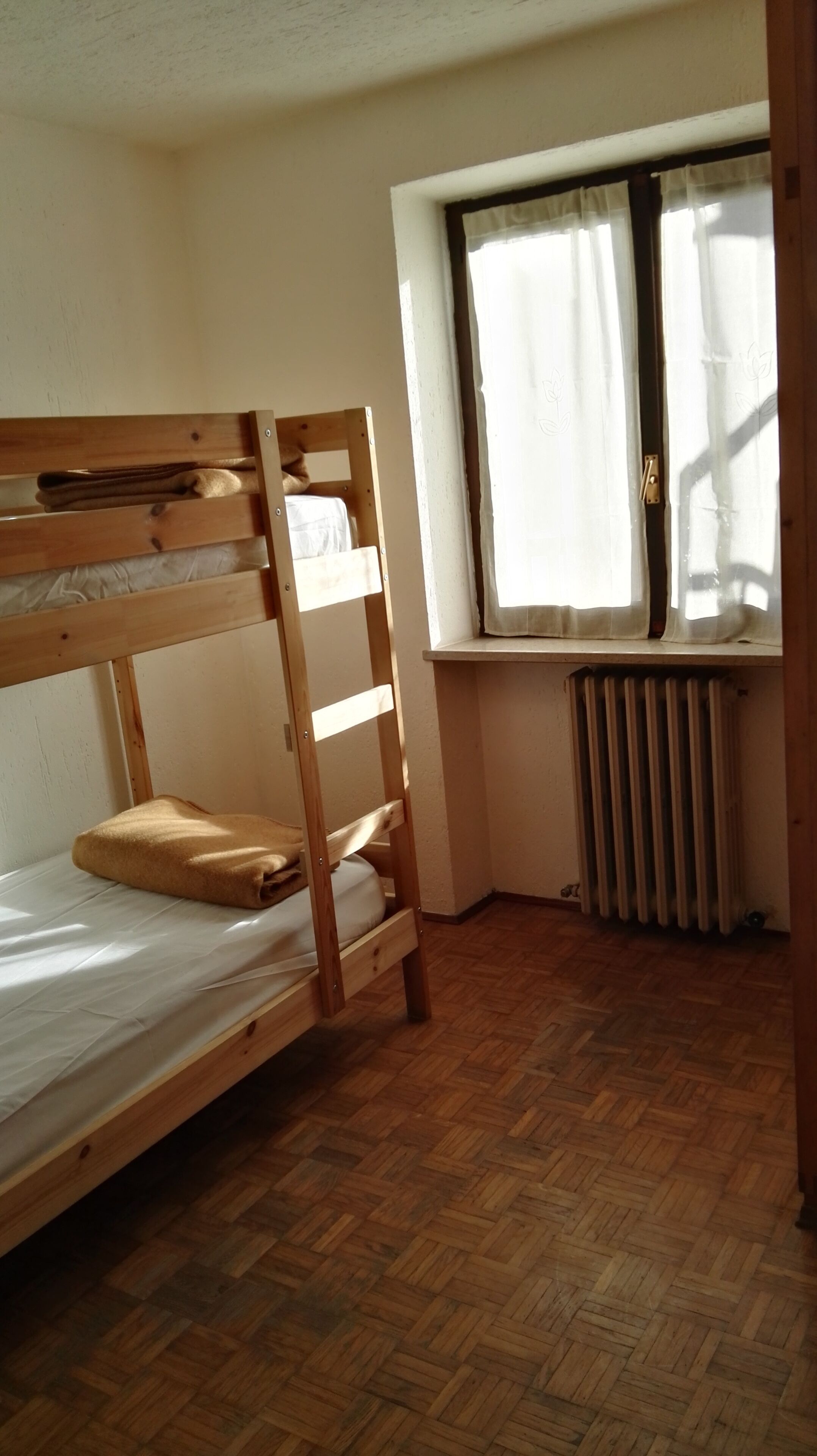 A bunk bed in a room with wooden flooring, a window with curtains, and a radiator.