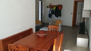En matplats med ett träbord och stolar, ett litet köksområde med ett kylskåp och en mikrovågsugn, och en hall som leder till ett annat rum.