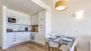A modern kitchen with white cabinets, a microwave, and a coffee maker. There is a dining area with a glass table and white chairs.