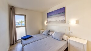 A bedroom with two beds, a large window with curtains, a painting of a beach scene, and bedside lamps.