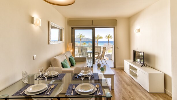 A modern living room with a glass dining table set for four, a sofa, a TV on a white cabinet, and a balcony with a view of palm trees and the sea.