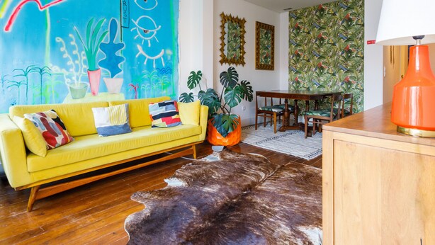 Un canapé jaune vif agrémenté de coussins colorés, une table à manger en bois avec des chaises, un tapis en peau de vache et une fresque murale éclatante.