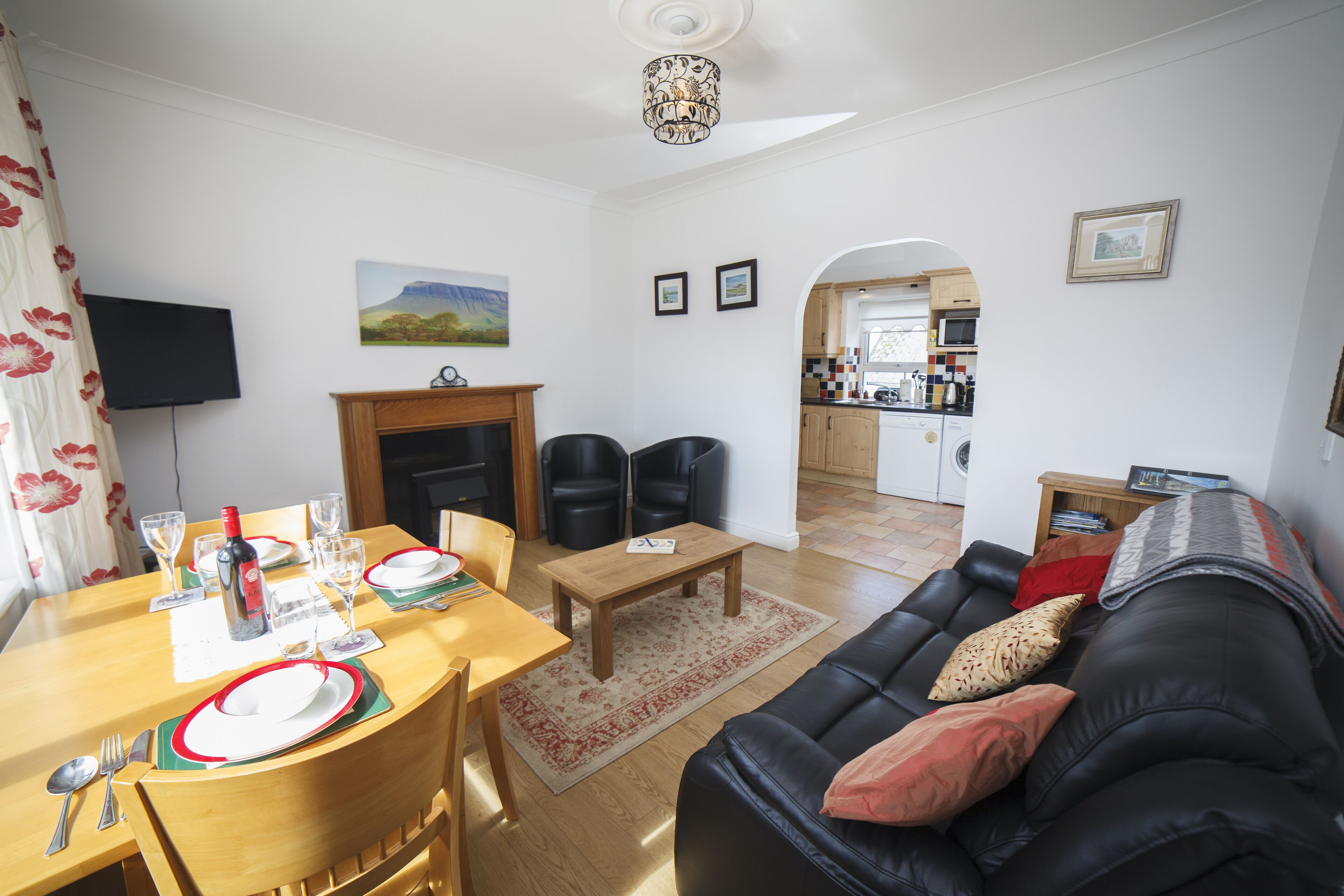 A living room with a black leather sofa, wooden dining table, and chairs. There is a fireplace, a television mounted on the wall, and a kitchen visible in the background.
