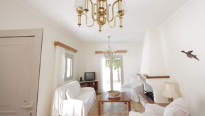 A spacious living room with a chandelier, white furniture, a television, and a view of the outdoors.
