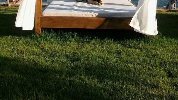 A wooden four-poster bed with white drapes, set on a grassy area with a view of a body of water and hills in the background.