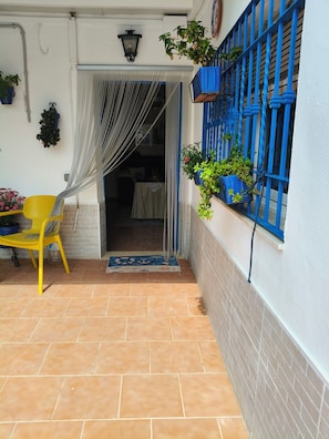 A covered patio with a yellow chair, a blue window with plants, and a white door with curtains.