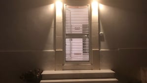 A building entrance with a metal door, illuminated by two wall-mounted lights, and a small potted plant to the left. - Sonetto holiday home in the center of Campomarino in Salento (Campomarino)