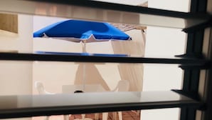A view through a window with a blue umbrella and white chairs, indicating an outdoor seating area. - Sonetto holiday home in the center of Campomarino in Salento (Campomarino)