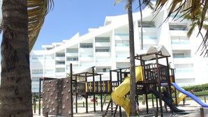 Um parquinho com escorregadores e estruturas de escalada, cercado por palmeiras e um edifício branco ao fundo.