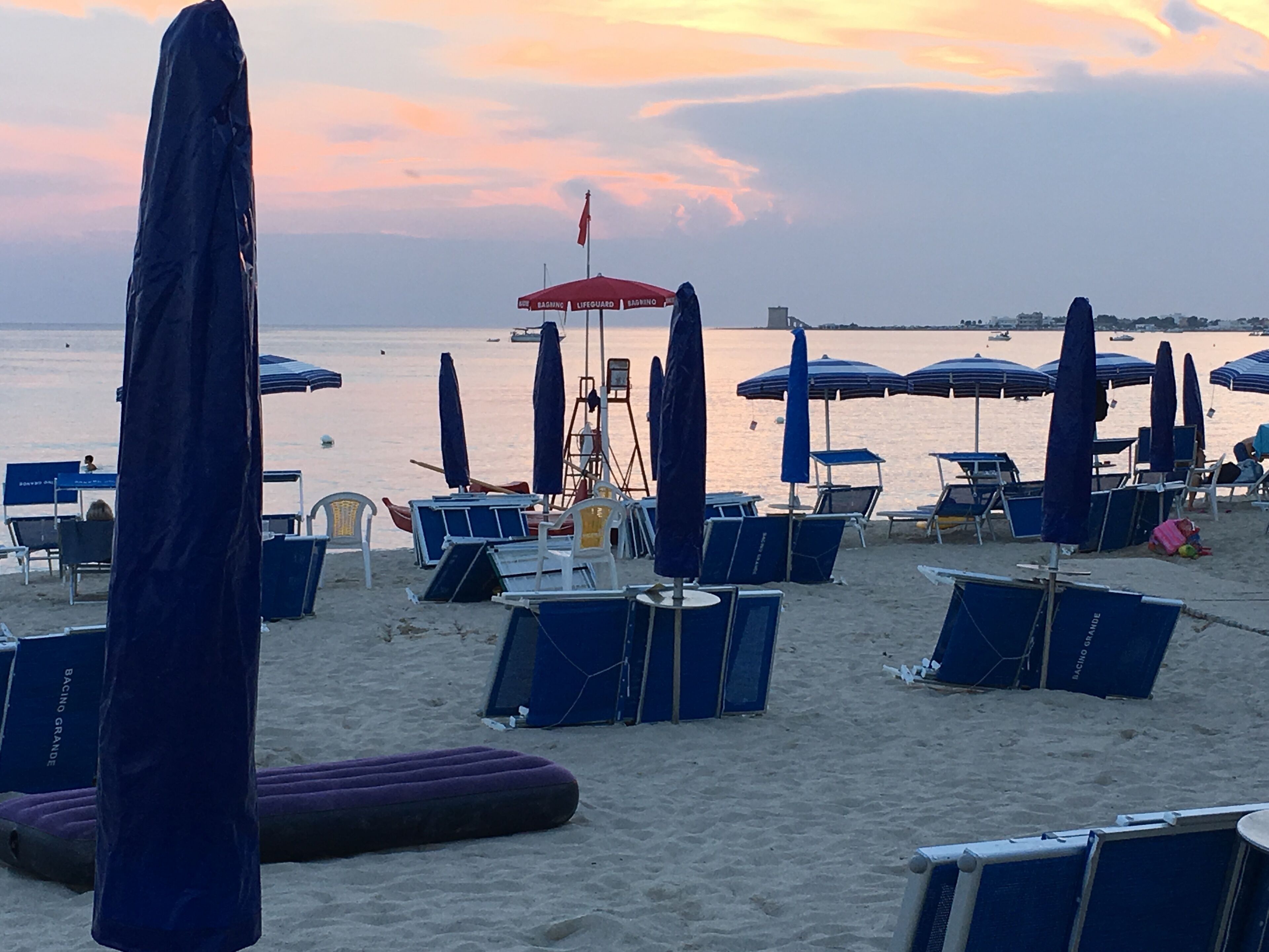 A beach with rows of blue lounge chairs and umbrellas, a lifeguard stand, and a calm sea in the background.