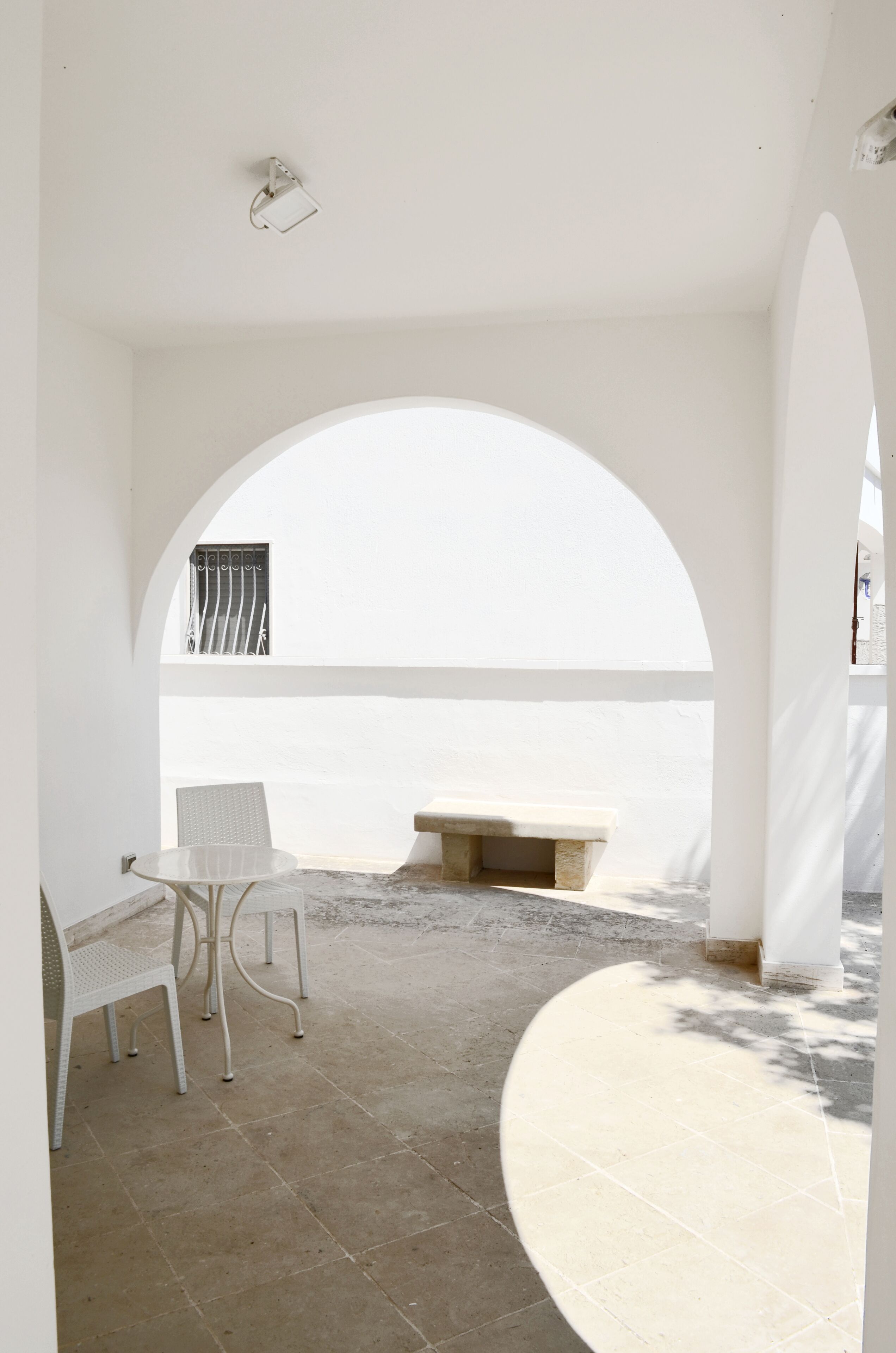 Een terras met uitzicht op zee, met een ronde tafel en twee stoelen, een stenen bank en een witte boog.