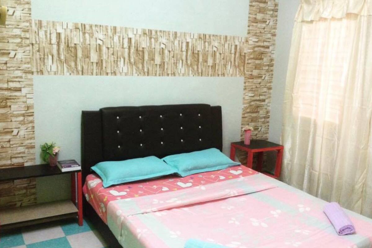 A bedroom with a bed, a nightstand, a red side table, a wall with a textured pattern, and a window with curtains.