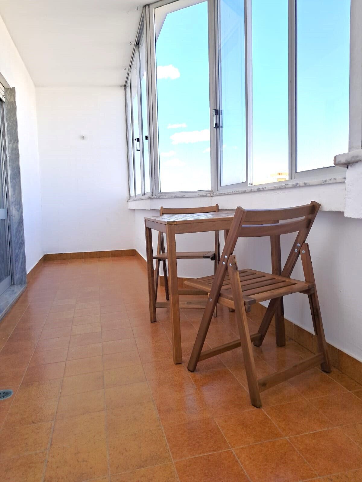 A room with a tiled floor, a wooden table and chair, and large windows with a view of the sky.