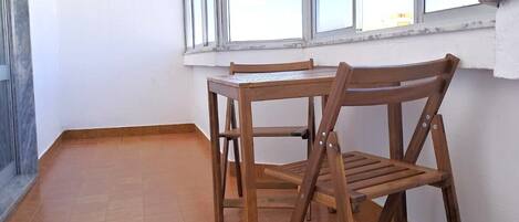 A room with a tiled floor, a wooden table and chair, and large windows with a view of the sky.