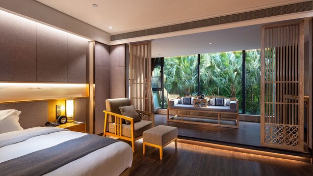 Una habitación de hotel moderna con una cama grande, una zona de estar con vistas a exuberante vegetación y un balcón con barandilla de madera.