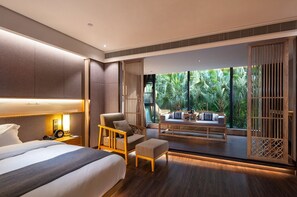A modern hotel room with a large bed, a seating area with a view of lush greenery, and a balcony with a wooden railing.