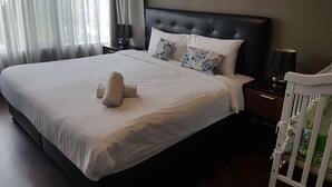 A neatly made bed with white linens and two pillows, a dark headboard, a bedside table with a lamp, and a crib with a blanket. - Baan San Dao Hua Hin (Hua Hin)