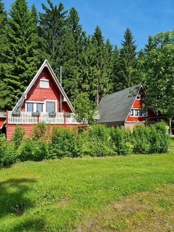 Deux cabanes rouges aux rambardes blanches, entourées d’arbres verdoyants et d’un champ herbeux.