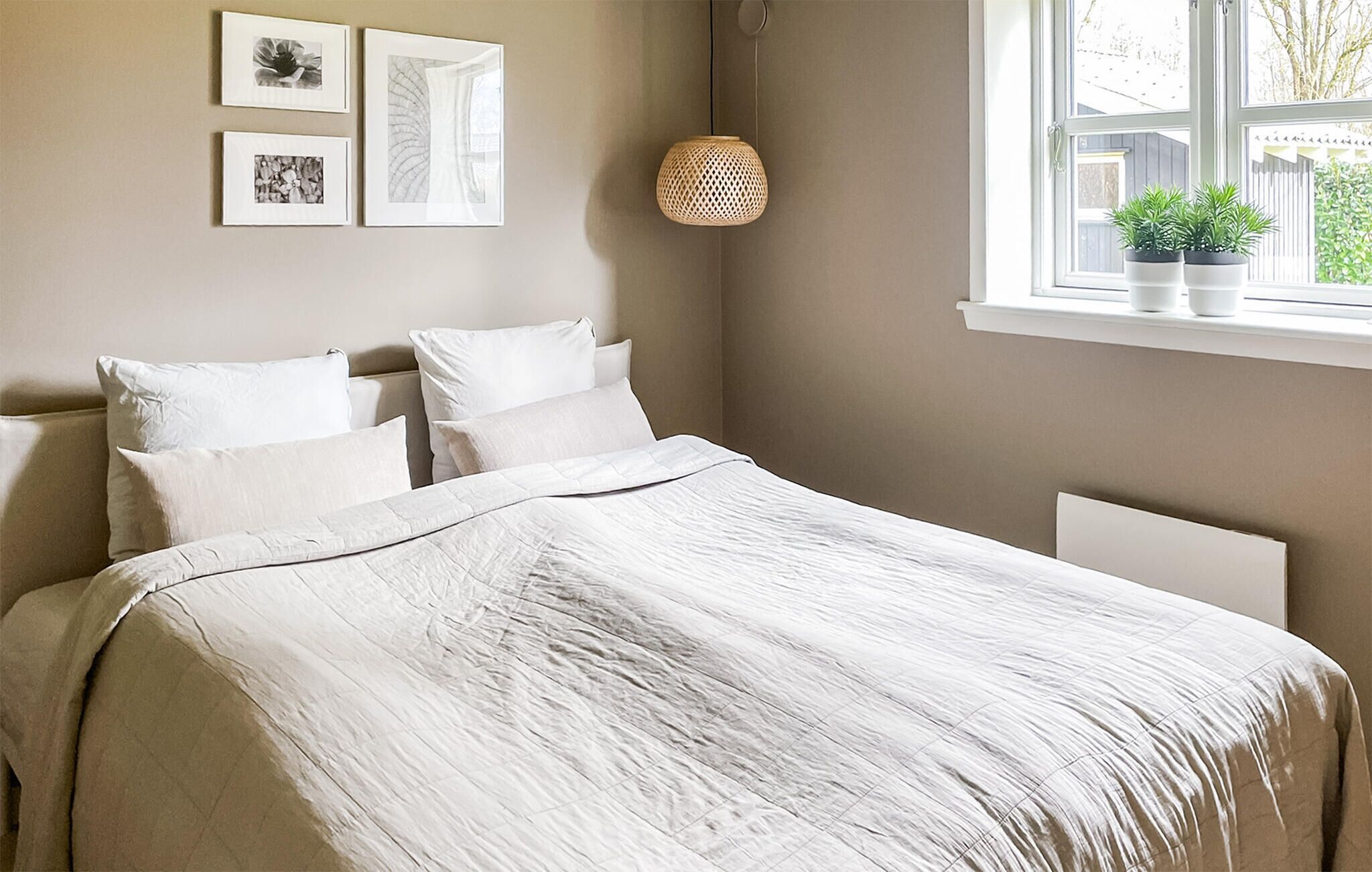 Une chambre à coucher avec un lit, des oreillers blancs, un luminaire suspendu et une fenêtre avec une plante.