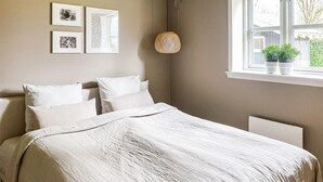 Ett sovrum med en säng, vita kuddar, en hängande lampa och ett fönster med en växt.